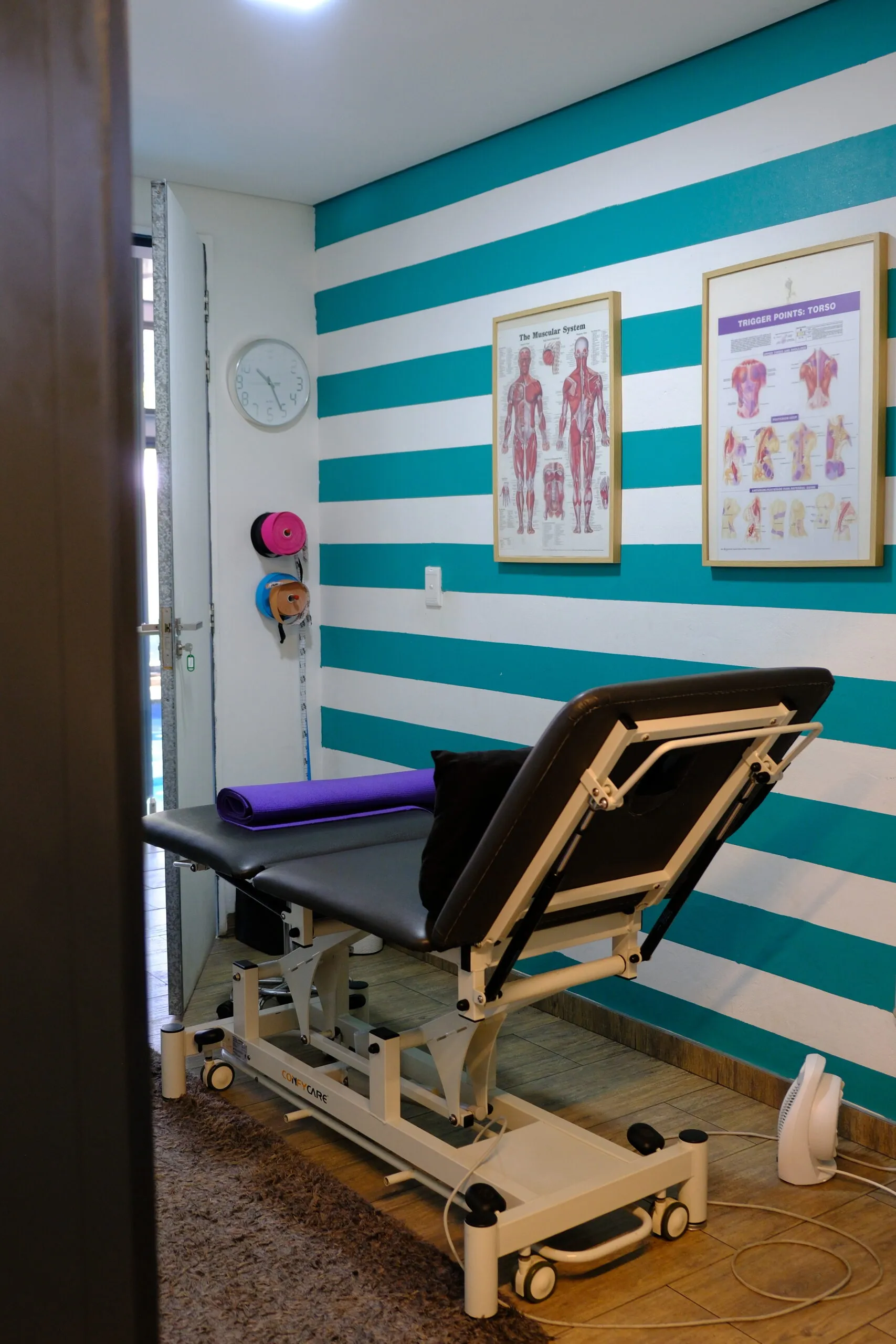 Adult treatment room with electric treatment table and anatomy references on the wall.